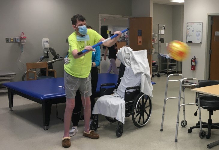 Johnny stands beside his wheelchair, holding a blue rod two-handed while a large, colorful ball flies toward him.
