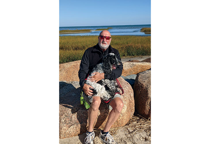 Paul sits on a rock with his cocker spaniel, Ellie, in his lap, and a marsh in the background.