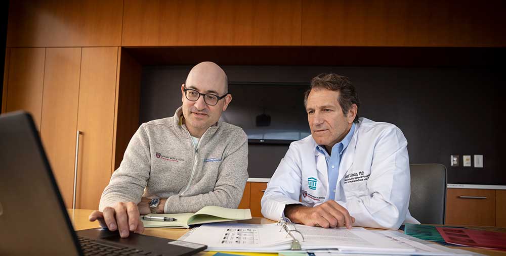 Two researchers consult a laptop at a paper-strewn desk.
